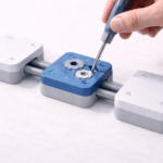 A hand uses a screwdriver to adjust a blue mechanical module connected between white rectangular components on a rail.